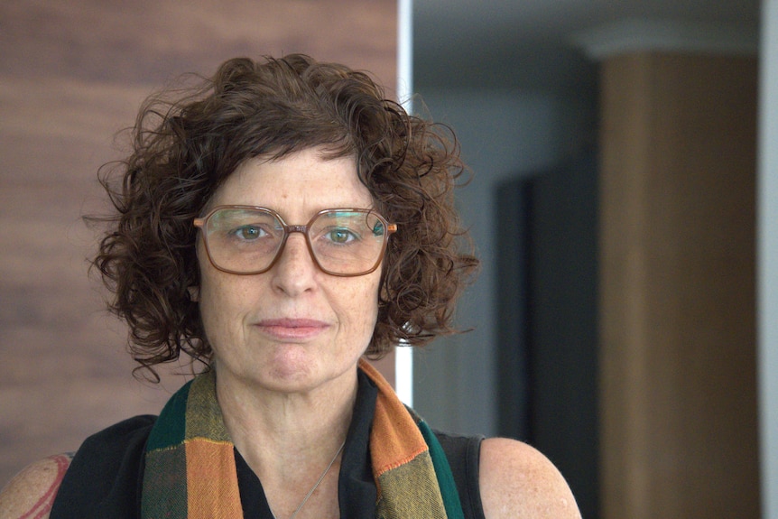 Portrait of a woman with short brown hair she is wearing glasses and a colourful scarf and looks straight ahead, non-smiling.