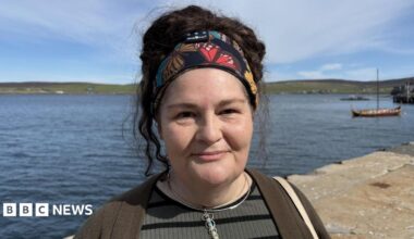 A 51-year-old woman with black hair looking at the camera and standing in a harbour area. Sea and hills are behind her and there's a small viking boat to the right. She is wearing a green top, necklace and her her is held up by a bandanna.