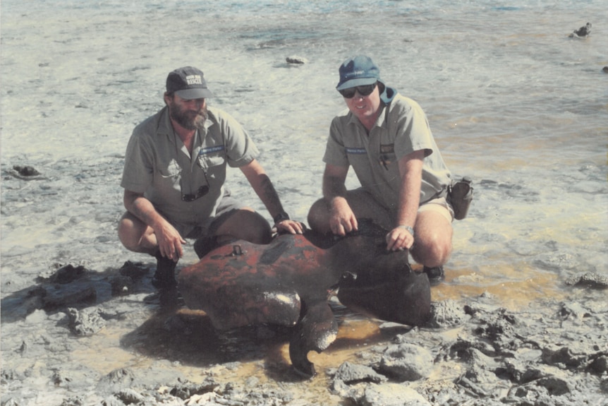 Two men wearing ranger gear kneel over an unexploded bomb on dusty ground.
