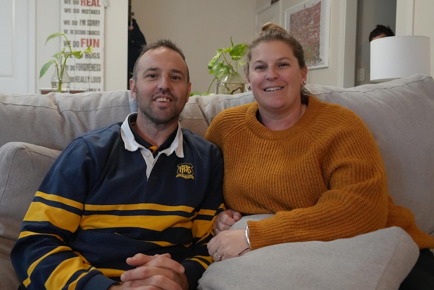 Daniel and Nicole Clark smile while sitting on a sofa, plants and table lamp behind.