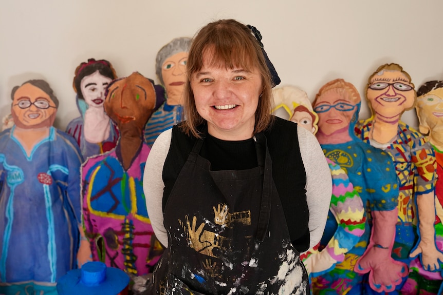 A woman smiles while standing in front of large people-like artworks.