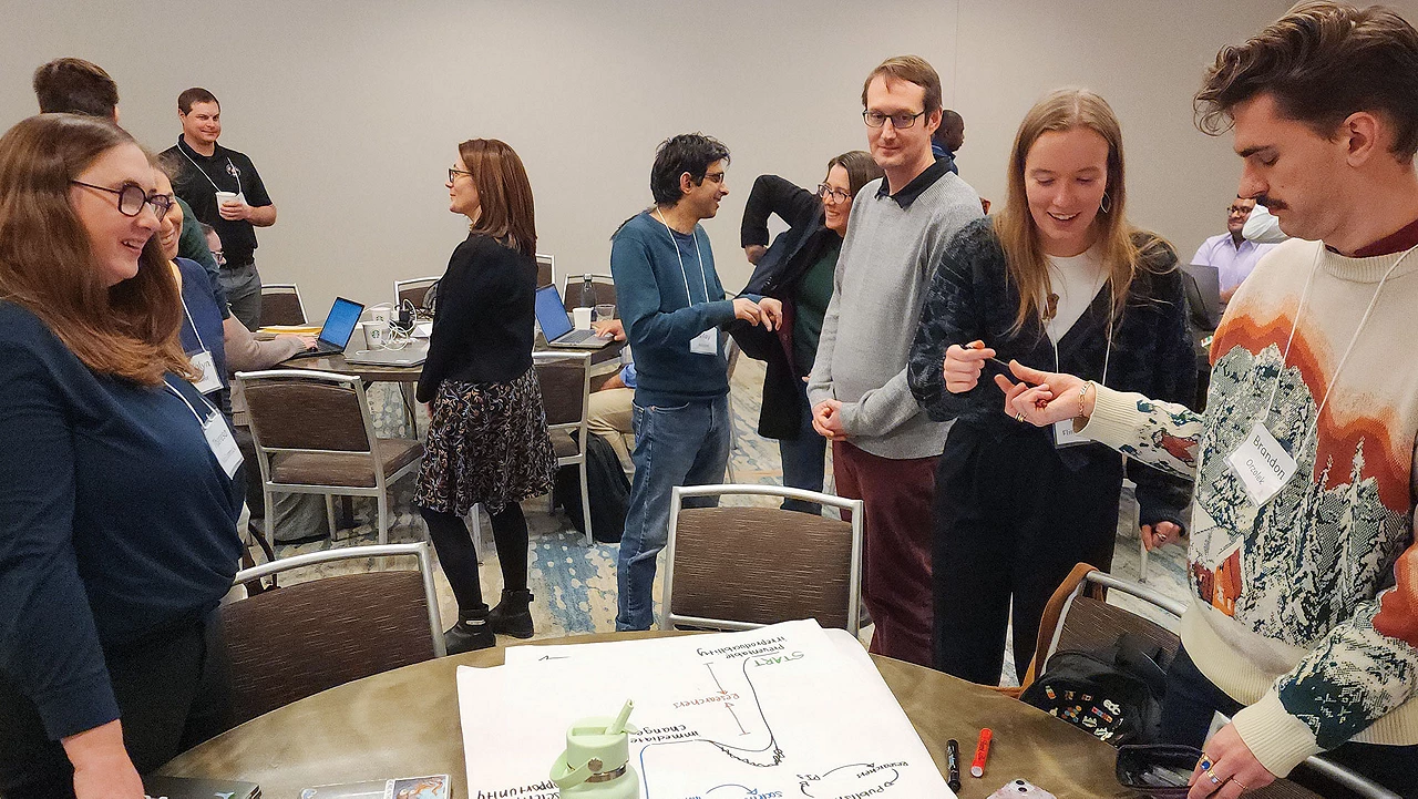 A group of people at a small event. A few are gathered around a table that holds a large diagram drawn on a white sheet of paper.