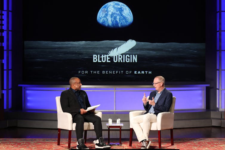 Two men sitting in chairs on a stage, with a screen behind them displaying the Blue Origin logo with a photo of the Earth from the Moon.