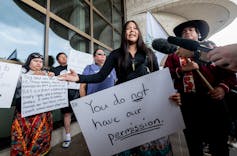 A young woman holding a placard, surrounded by other protesters, speaks into microphones from various media outlets