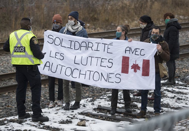 A police officer talks to protesters holding a banner across the railway tracks bearing the words ‘Solidarity with Indigenous struggles.'