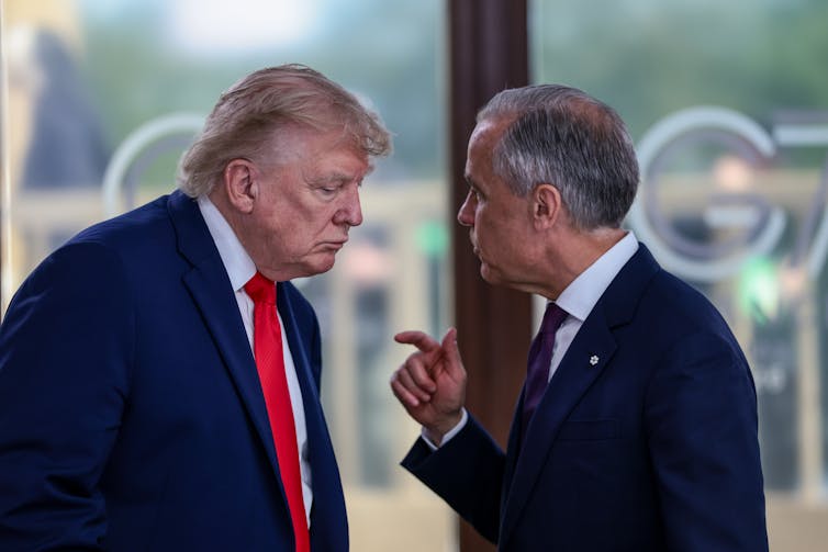 Two white men in dark suits stand next to each other speaking and gesturing