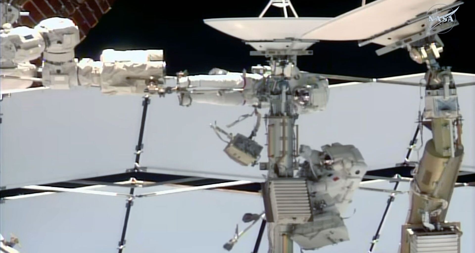 two astronauts dressed in white spacesuits work near a couple of antenna dishes outside of a space station.