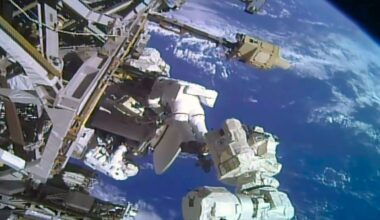 two astronauts in white spacesuits work near a large antenna dish outside a space station with a blue and white planet as their backdrop