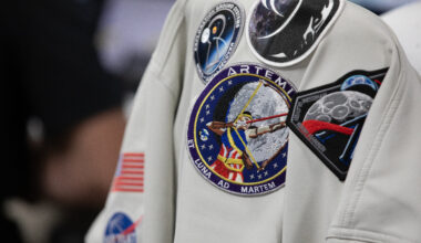 A gray colored jacket with five Artemis mission patches and a NASA insignia hangs on a chair.