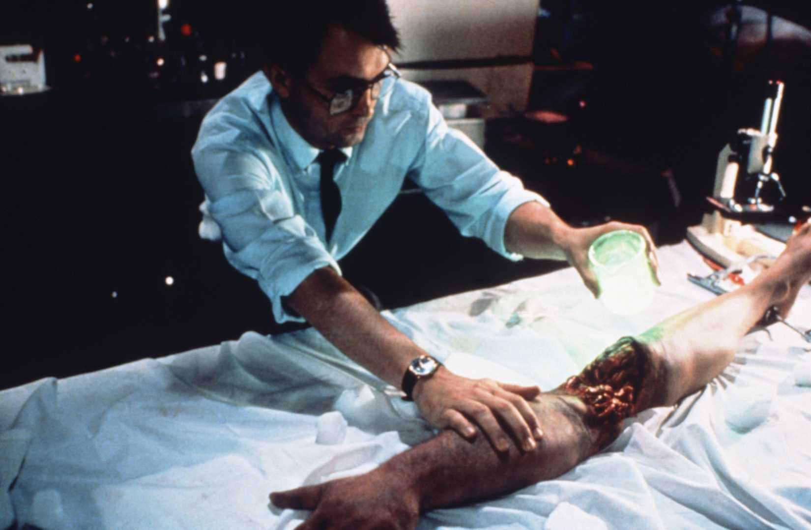 A bespectacled Herbert West uses his neon reagent on a severed arm and leg joined together in Bride of Re-Animator