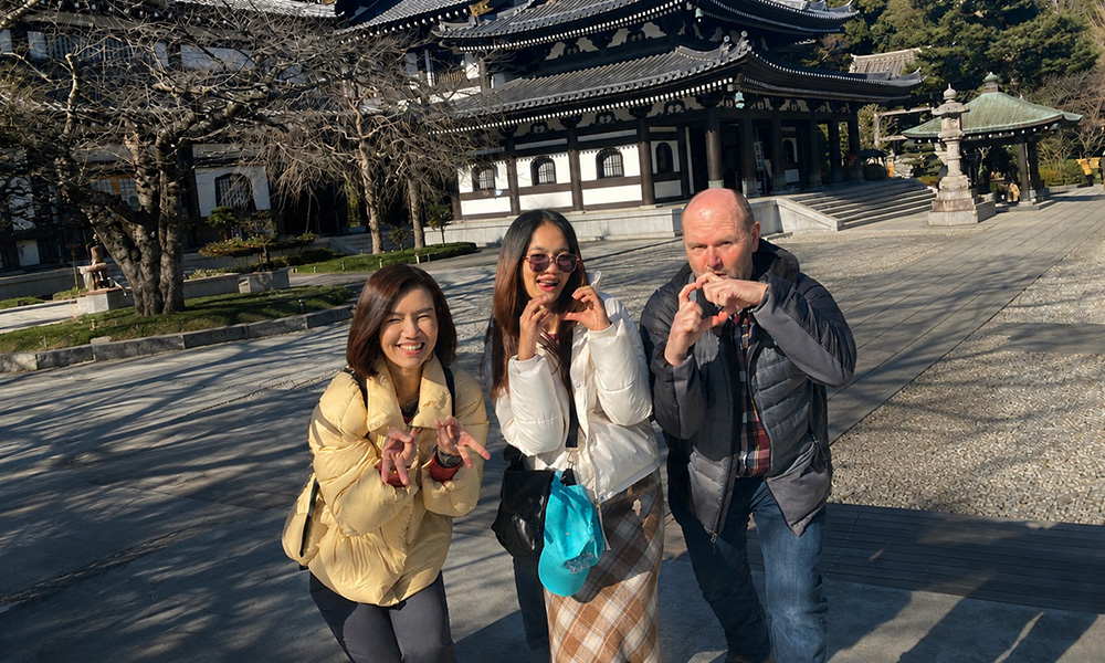Three people pose comically for a photo outside an ornate-looking Japanese museum, with full green trees framing the photo.