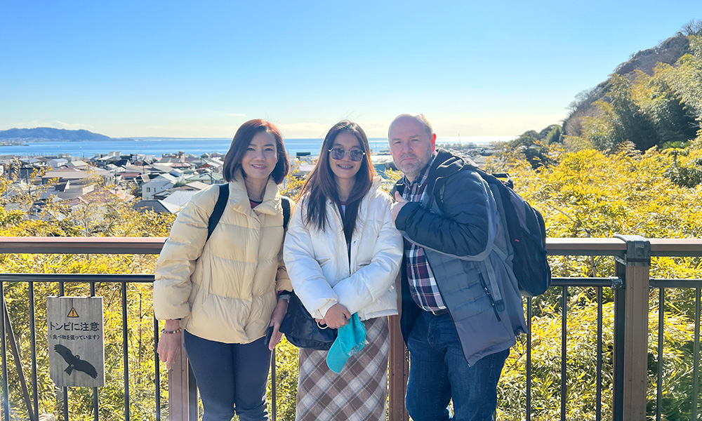 Three people-Siriporn on the left, Pimjai Pimbaotham in the centre, and Robert on the right-pose together at a railed lookout on a sunny day. All three wear jackets and backpacks. Behind them are a coastal town, the sea, and autumn-coloured hillsides, with a small sign in Japanese mounted on the railing at left.