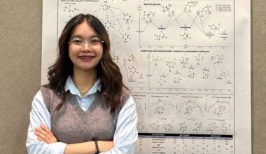 A young woman with long dark hair and glasses, wearing a grey knit vest over a light blue collared shirt, smiles with her arms crossed in front of a large research poster covered in molecular diagrams and energy graphs titled