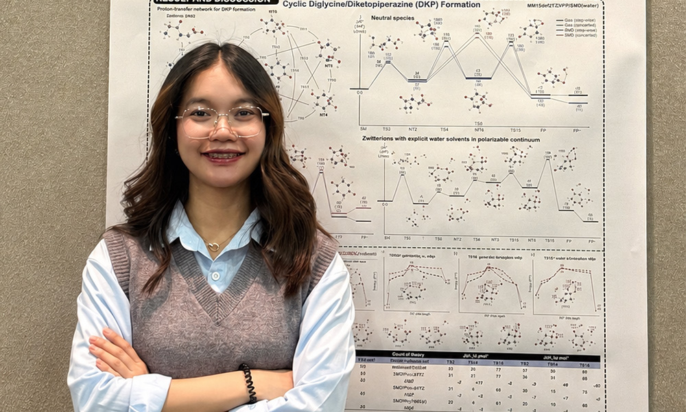 A young woman with long dark hair and glasses, wearing a grey knit vest over a light blue collared shirt, smiles with her arms crossed in front of a large research poster covered in molecular diagrams and energy graphs titled