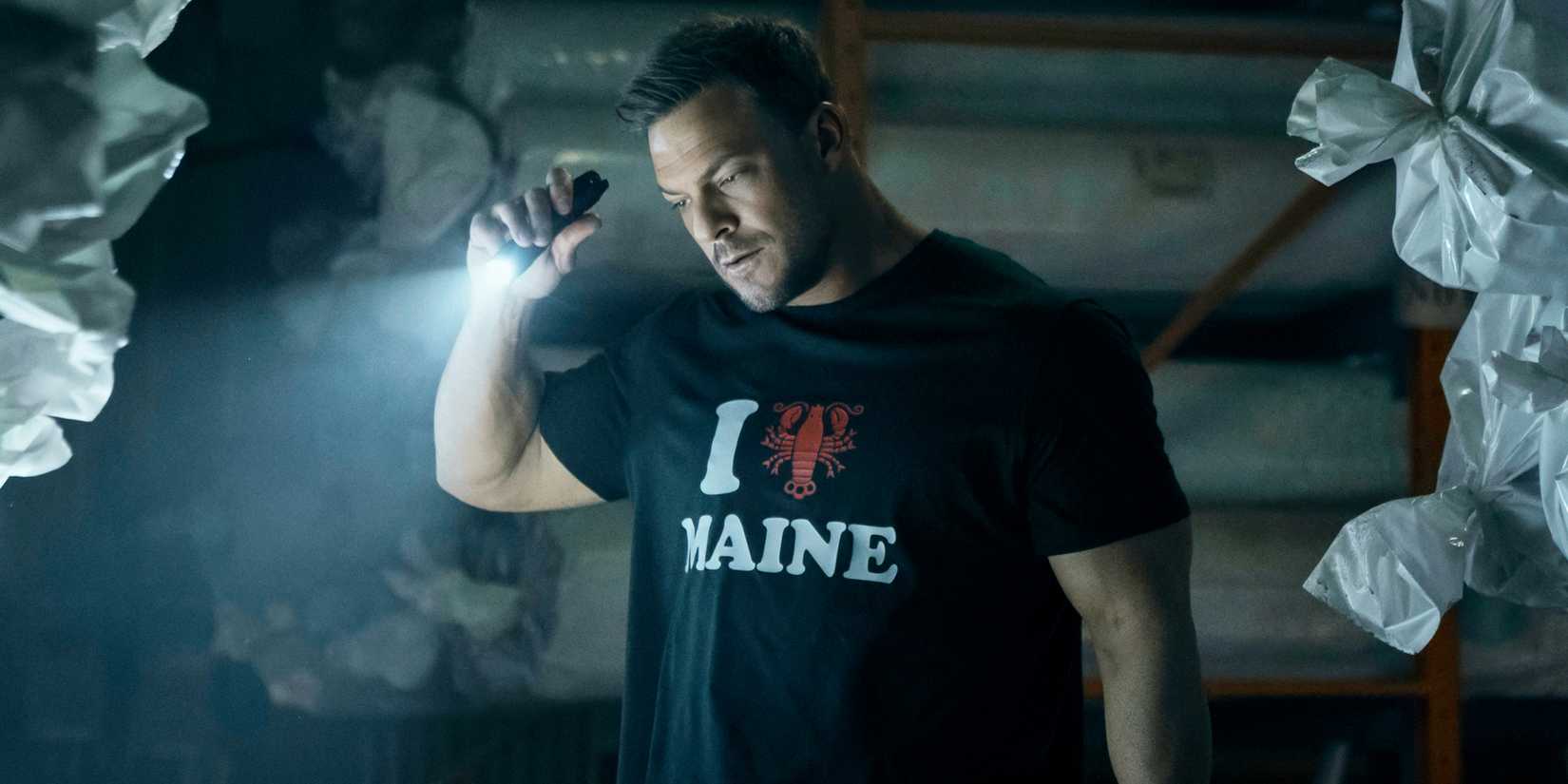 Alan Ritchson holds a flashlight and wears a Maine novelty shirt in Reacher.
