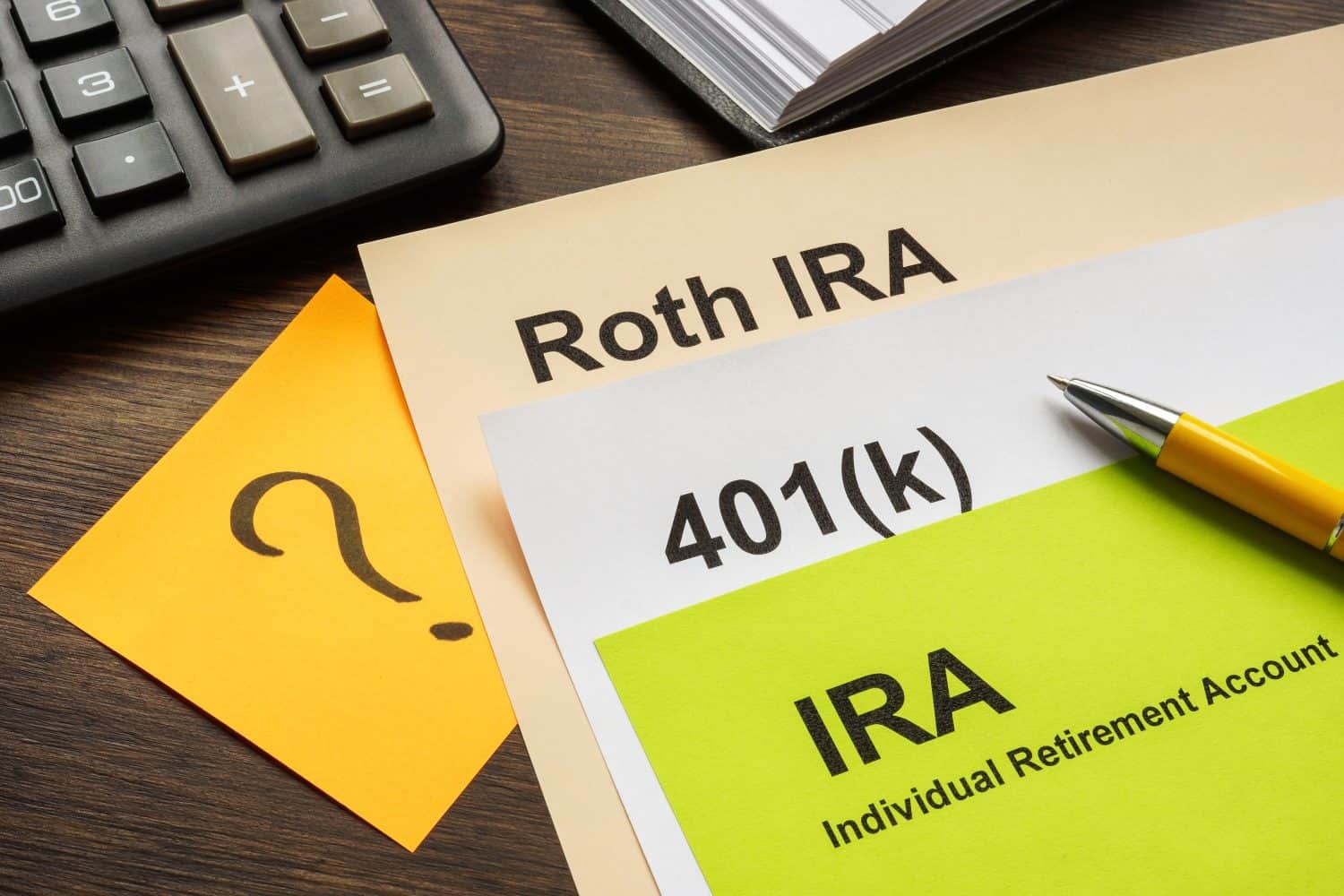 A flat lay image on a dark wood desk featuring beige and white financial documents. The top document is labeled 'Roth IRA', the middle '401(k)', and the bottom 'IRA' with 'Individual Retirement Account' underneath. A bright yellow sticky note with a large black question mark covers part of the left side. A black calculator is partially visible in the upper left, and a silver and yellow pen rests on the 'IRA' document on the right.