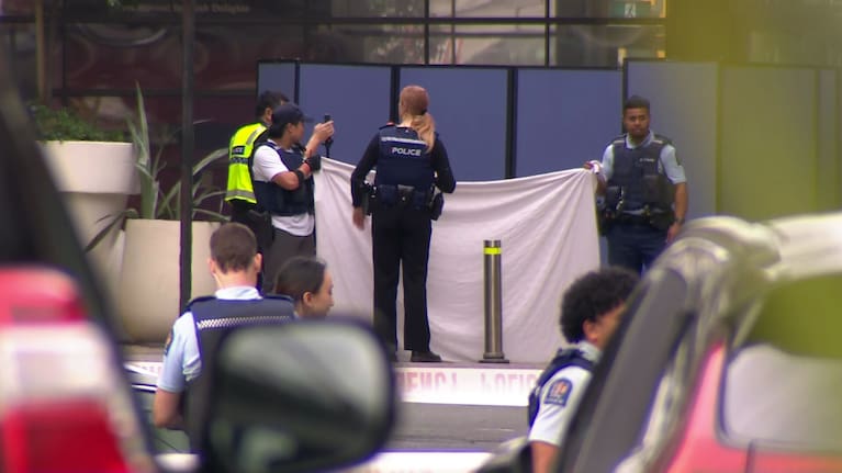 Police beyond a cordon at Sylvia Park holding up a white sheet to shield part of the scene from public view.