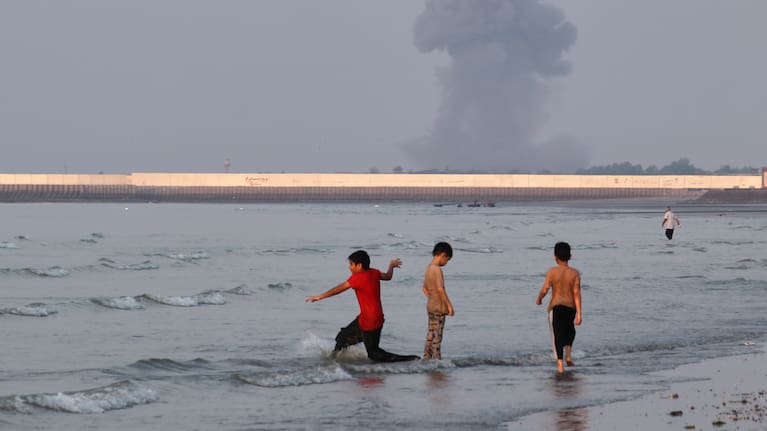 Three boys play in the shallow waters of the Strait of Hormuz, as a plume of smoke rises from an explosion in the background, off Bandar Abbas, Iran, Monday, July 13, 2026