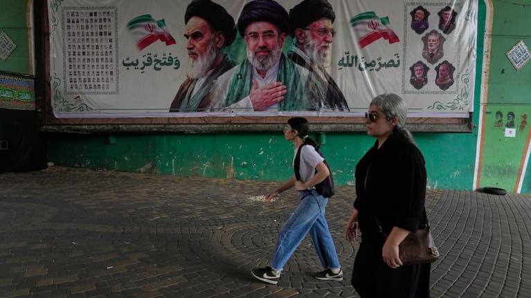 Women walk past a banner showing portraits of the late Iranian revolutionary founder Ayatollah Khomeini, left, Supreme Leader Ayatollah Mojtaba Khamenei.