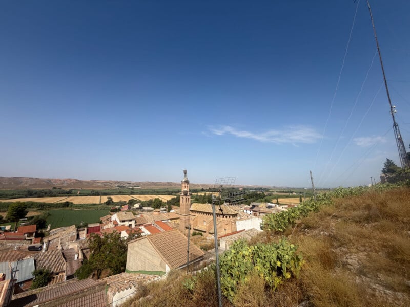 A small village with a prominent church tower sits nestled in a valley surrounded by agricultural fields under a clear blue sky.