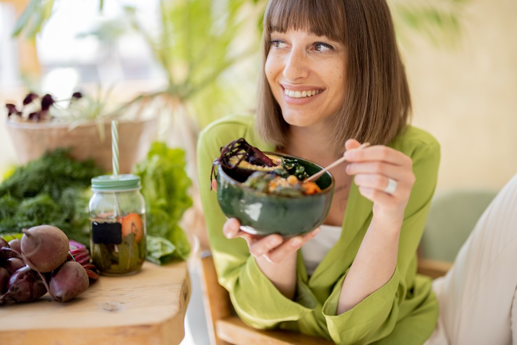 A cheerful young woman with a bob haircut smiles while eating a vegetarian lunch from a green bowl.