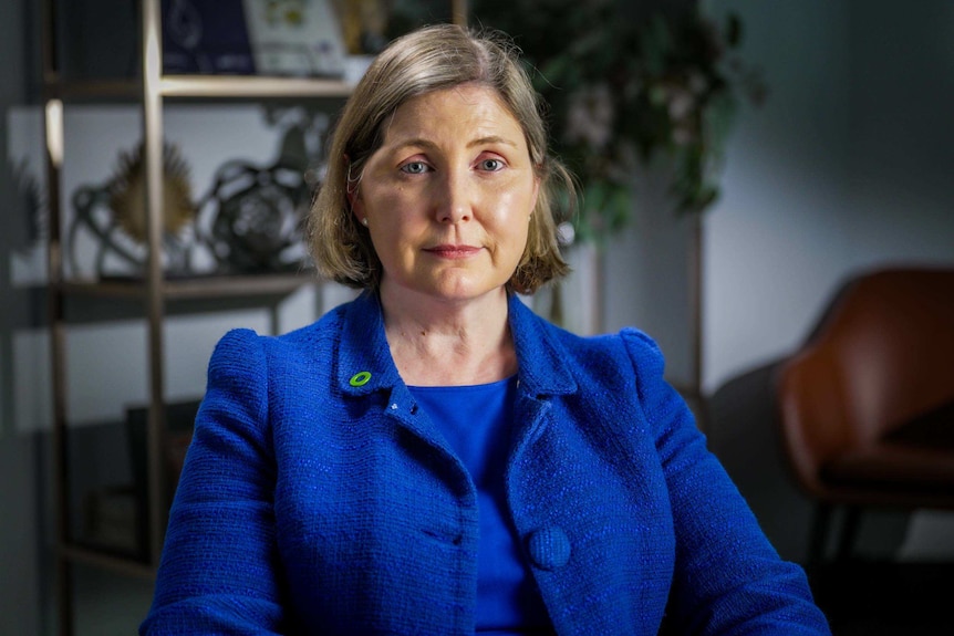 A blonde-haired woman sits in an office weaing a blue jacket.