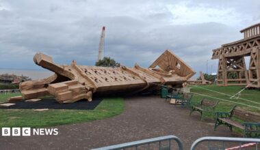 A collapsed cardboard structure on its side in a park laying on top of grass and a path. To the right, a standing piece of the artwork still stands. There is a crane behind and to the left you can see the Bristol Channel in the background along with a small section of Birnbeck Pier. In the park there are iron framed wooden benches which are faded green in colour.