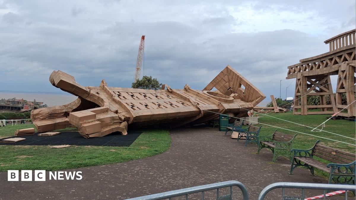 A collapsed cardboard structure on its side in a park laying on top of grass and a path. To the right, a standing piece of the artwork still stands. There is a crane behind and to the left you can see the Bristol Channel in the background along with a small section of Birnbeck Pier. In the park there are iron framed wooden benches which are faded green in colour.