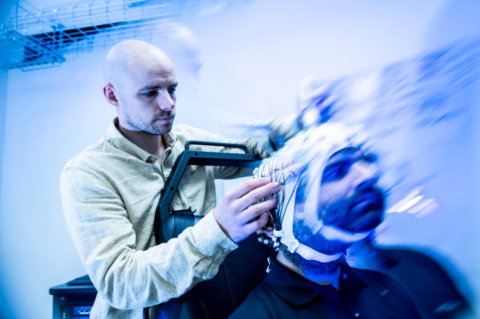 A motion-blurred, blue-toned shot of Sherman adjusting electrodes on a participant's cap, with wires trailing behind the head in a streak of movement.