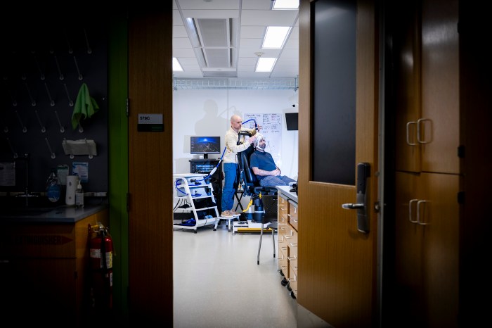 Sherman fits a participant with an EEG cap in the lab, viewed through an open doorway with equipment carts and a wall-mounted monitor visible in the foreground room.
