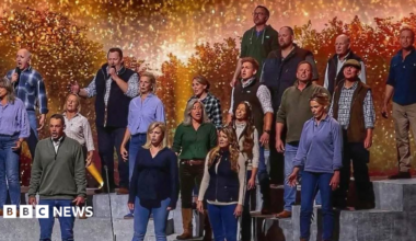 The choir are standing on tiered steps on a stage. They are all wearing rugged farmers' clothing of jeans, checked shirts, gilets and hoodies in shades of blue, purple and green. There are sparkling orange lights twinkling in the background