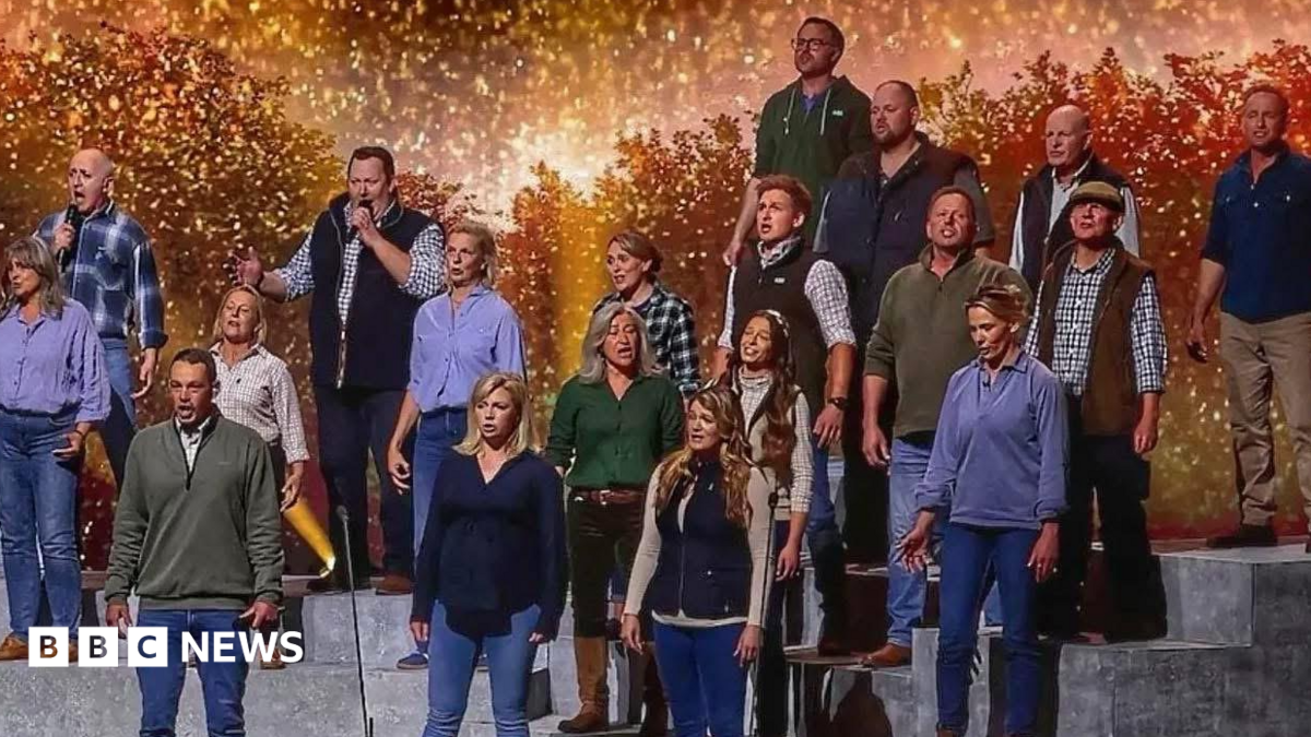 The choir are standing on tiered steps on a stage. They are all wearing rugged farmers' clothing of jeans, checked shirts, gilets and hoodies in shades of blue, purple and green. There are sparkling orange lights twinkling in the background