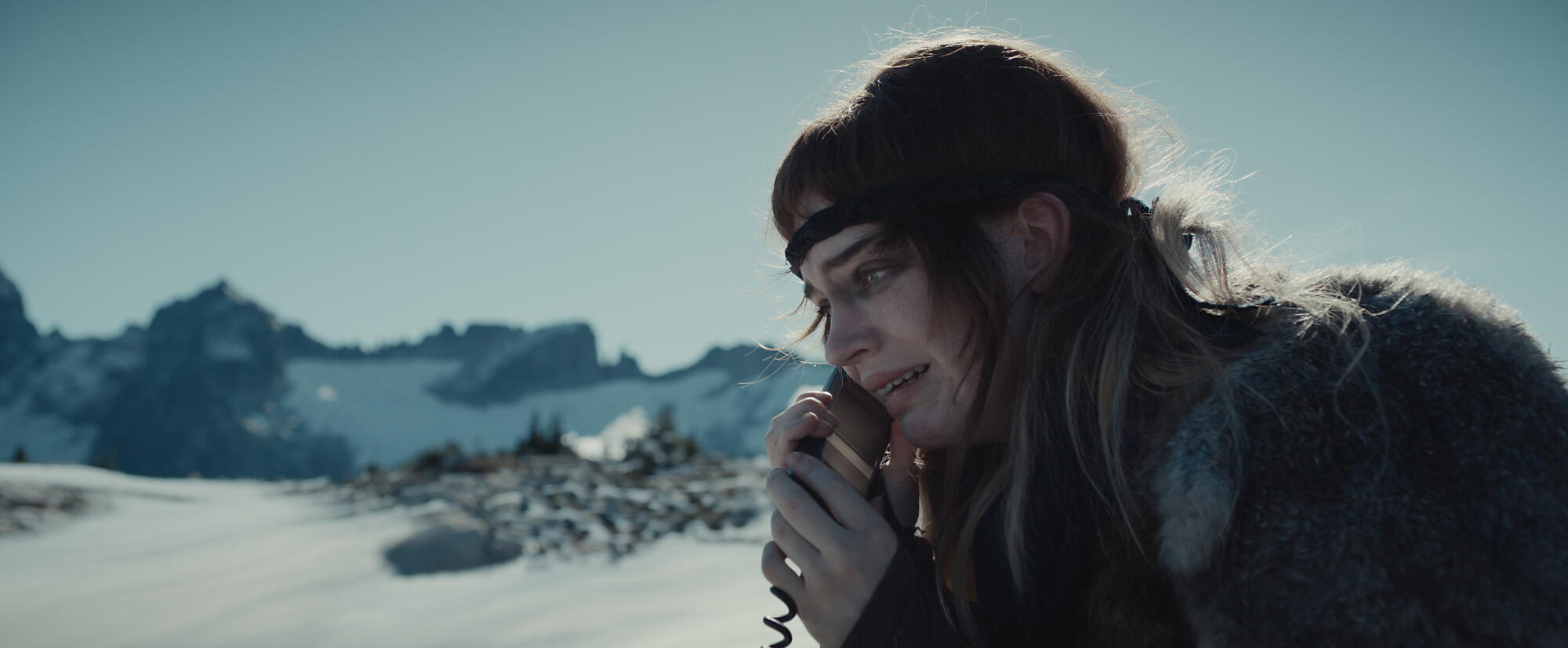 A teenage girl with a frightened expression holds a satellite phone to her ear on a snowy mountaintop.