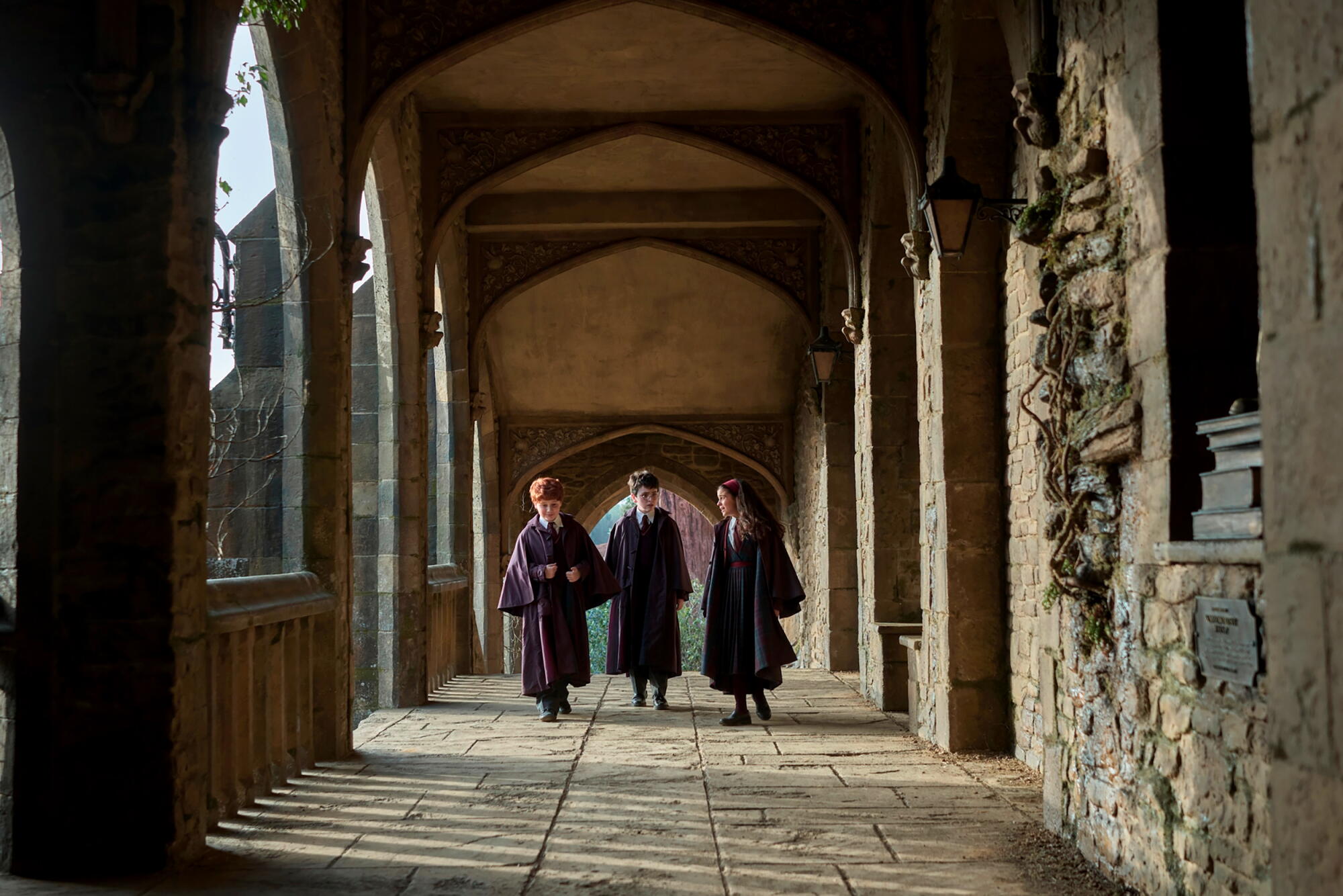 Two boys and a girl in robes walk down a covered stone walkway.