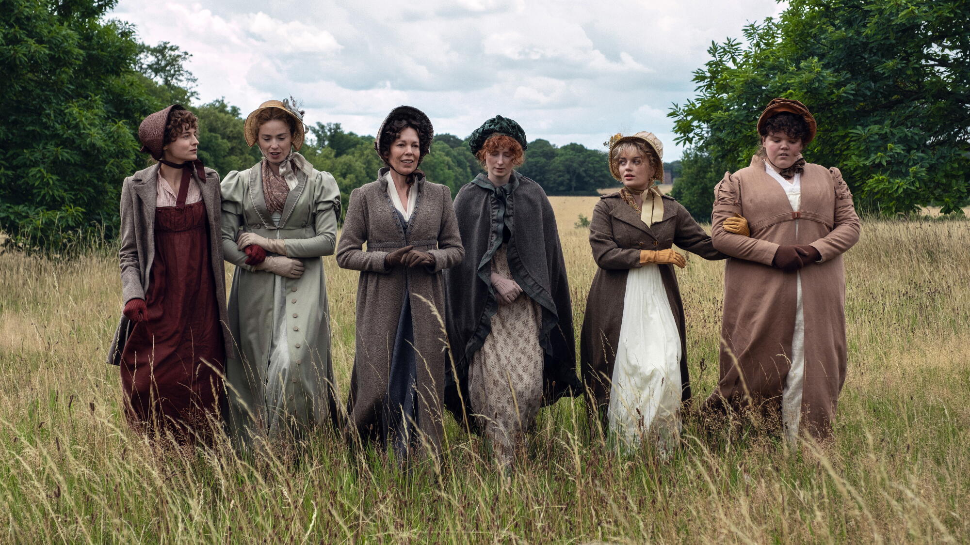 Six women wearing bonnets, long dresses and gloves walk through an overgrown field.