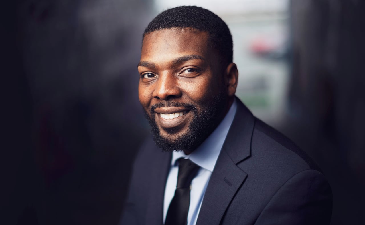 A man with black hair, beard and moustache wearing a dark blue business suit smiles.
