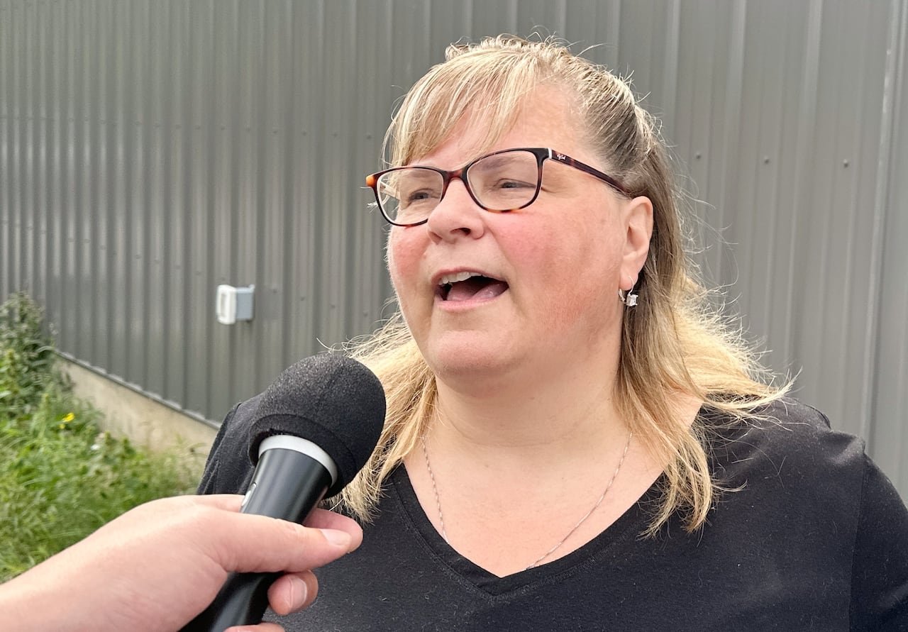 A woman with long blonde hair and glasses wearing a black t-shirt speaks into a microphone.