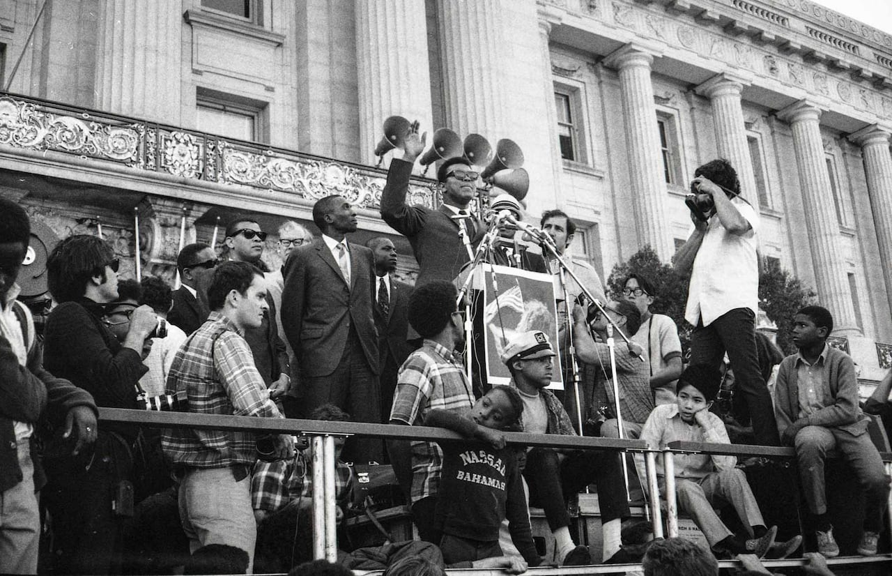 A black and white photo shows a Black man in black sunglasses on a stage with one arm raised, surrounded by people sitting and standing around him and a building with white columns behind him.
