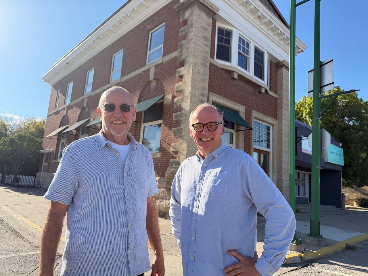 Two men in pale blue shirts stand outside on a street in front of a historic brick building.