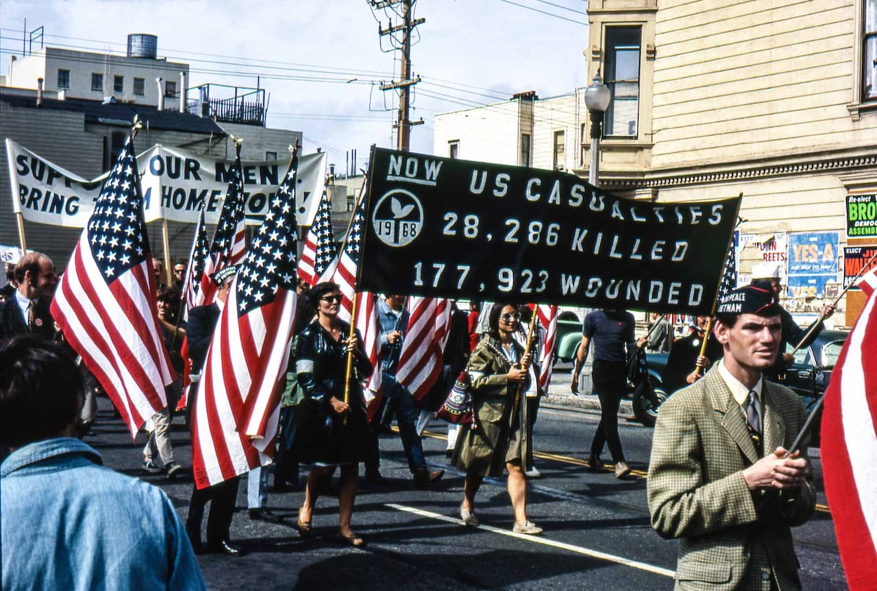 People holding American flags and signs with statistics on people killed and wounded march through a street.