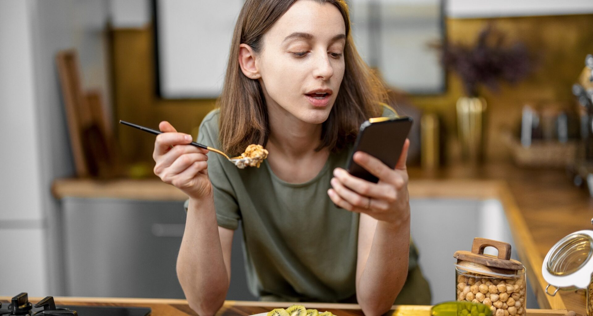 Woman in casual green t-shirt holds a spoon with granola in her hand and reads on the phone during a morning time on the kitchen at home.