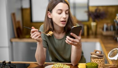 Woman in casual green t-shirt holds a spoon with granola in her hand and reads on the phone during a morning time on the kitchen at home.