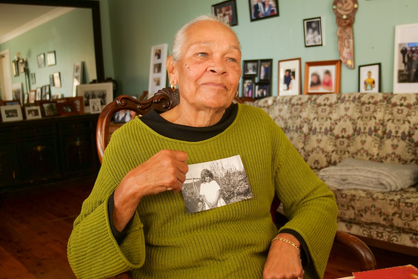 Ms Cruse-Rawiri sitting in a chair looking into the distance while holding a black and white photograph of herself smiling.