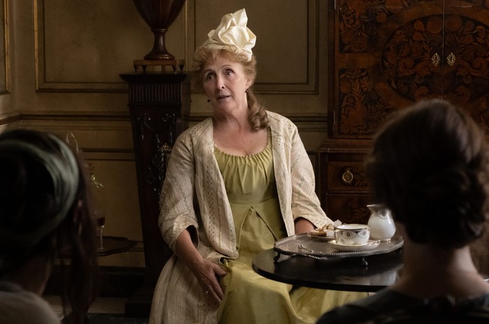Mrs Jennings (Fiona Shaw) in the new adaptation. Jennings is a financially independent woman – contradicting many stereotypes about women of the period | Credit: © 2026 FOCUS FEATURES Film still of an older woman in a period drama, seated in an ornate wood-paneled room. She wears a pale yellow-green dress with a striped cream shawl and a tall cream bow headpiece over curled hair, speaking with an animated expression. In front of her is a small round table with a tea set — cup, saucer, creamer, and a plate of biscuits — on a silver tray. Two out-of-focus figures are visible in the foreground