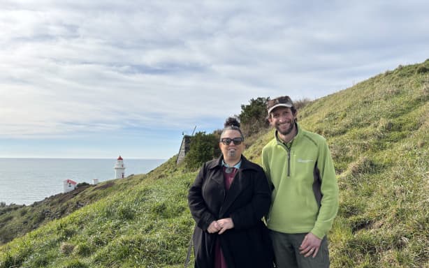 Nadia and Jim stand in the long grass on the headland. Behind them in the midground is an overgrown stone structure, further in the background a lighthouse is visible. It's partially cloudy, they are both smiling at the camera.