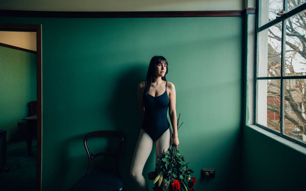 Amy is wearing a black leotard and standing against a green wall. to her right is a big window which she is gazing out of. In her left hand she is holsing a very leafy bunch of red roses.