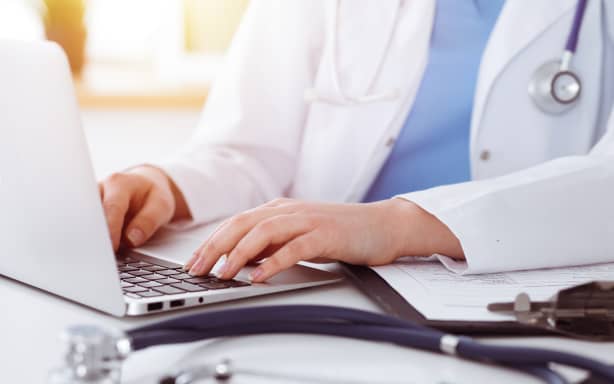 Unknown woman-doctor typing on laptop computer while sitting at the table in sunny clinic.