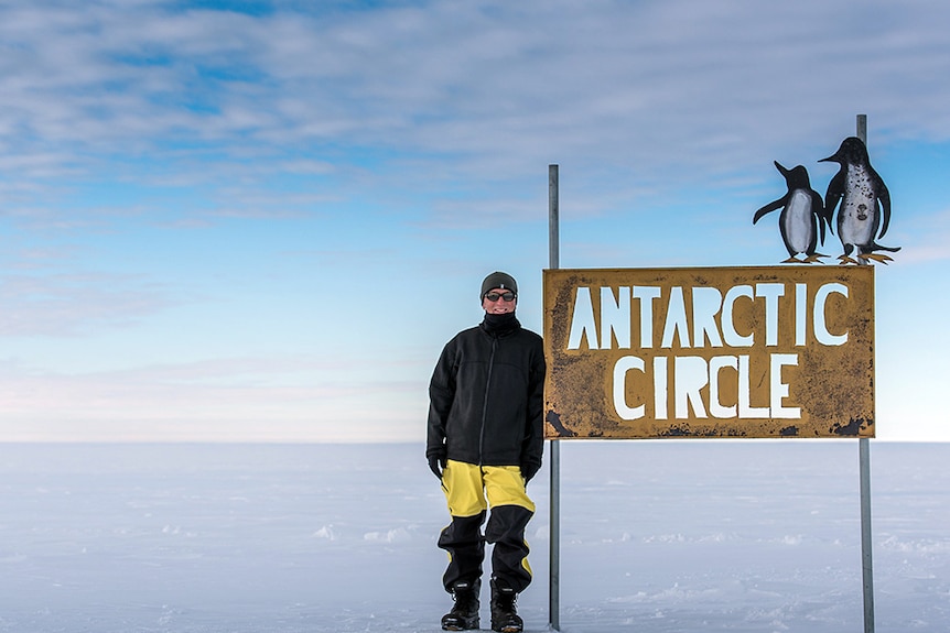 Nick Gales stands next to a sign in Antarctica that reads Antarctic Circle.