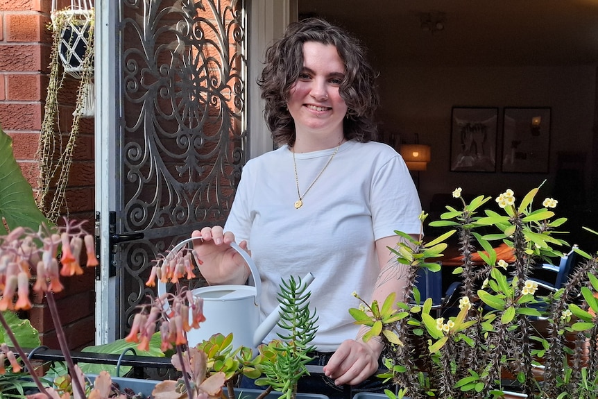 A young woman on a blacony with lots of plants, smiling and holding a watering can