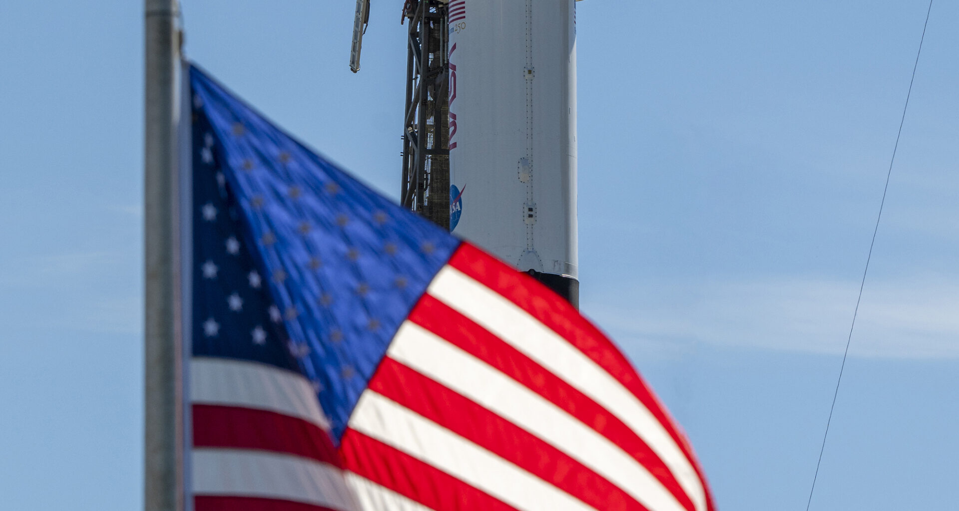 A close-up view of a SpaceX Crew Dragon spacecraft mounted atop a Falcon 9 rocket on the launch pad. The capsule and trunk are connected to ground support hardware, with NASA and mission insignia visible on the rocket’s exterior against a bright blue sky. in the foreground in the American flag.